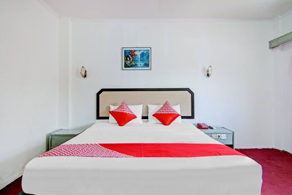 Un lit bien fait, recouvert d’une couvre-lit rouge et blanche, avec deux oreillers rouges et une petite table de chevet sur laquelle se trouvent un téléphone et une prise électrique.