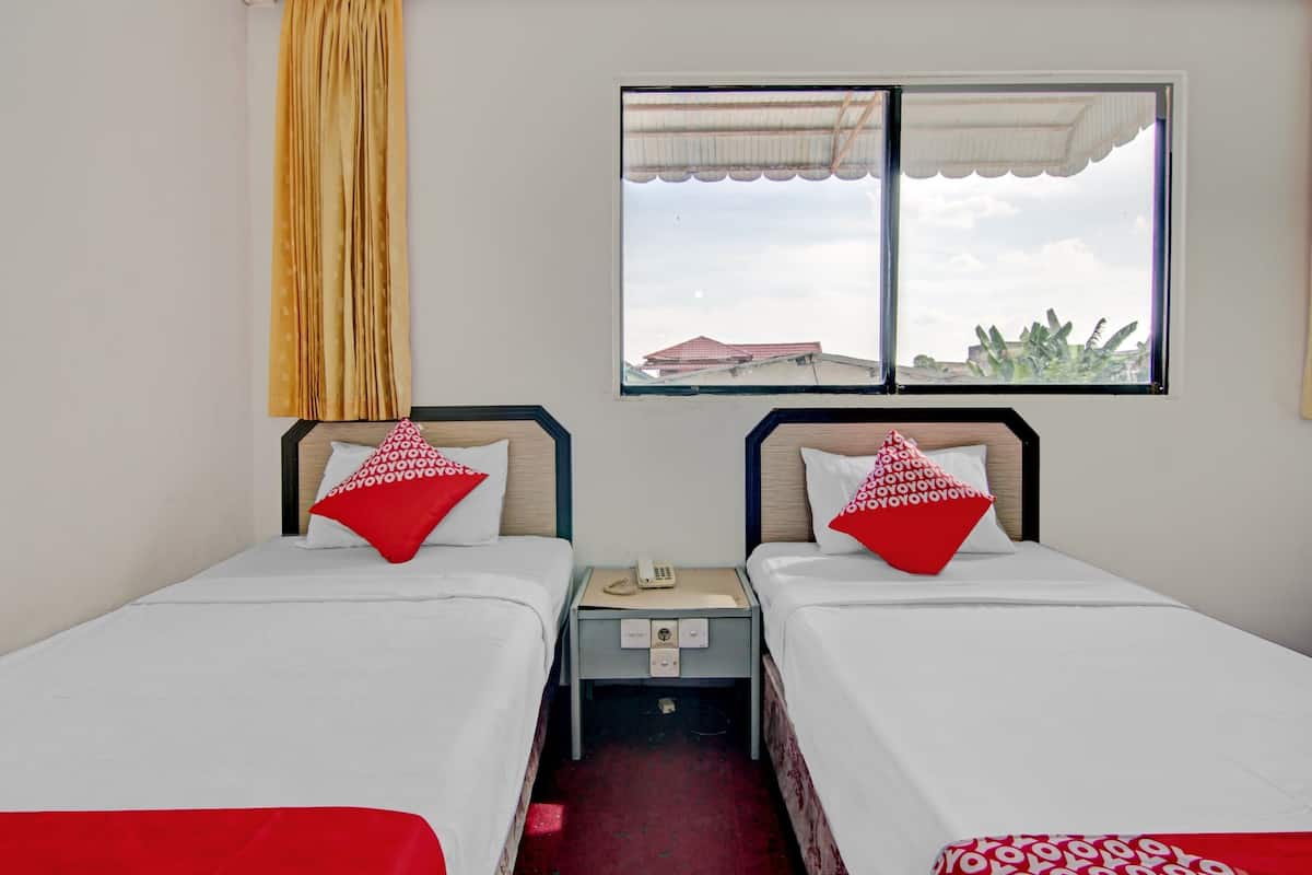 Une chambre d’hôtel avec deux lits simples, chacun agrémenté d’un oreiller rouge et d’une couverture blanche. On y trouve une petite table de chevet avec un téléphone et une étagère fixée au mur. Une fenêtre offre une vue sur des immeubles et un bananier.
