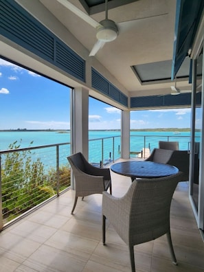 Ein Balkon mit rundem Tisch und Stühlen, der Blick auf ein türkisfarbenes Meer und das entfernte Land gewährt.