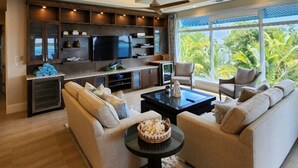 Un salon moderne doté d’un grand téléviseur, de sièges confortables et offrant une vue sur une végétation luxuriante.