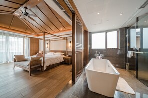 A modern hotel room with a large bed, a separate sitting area, a bathtub, and a glass shower.