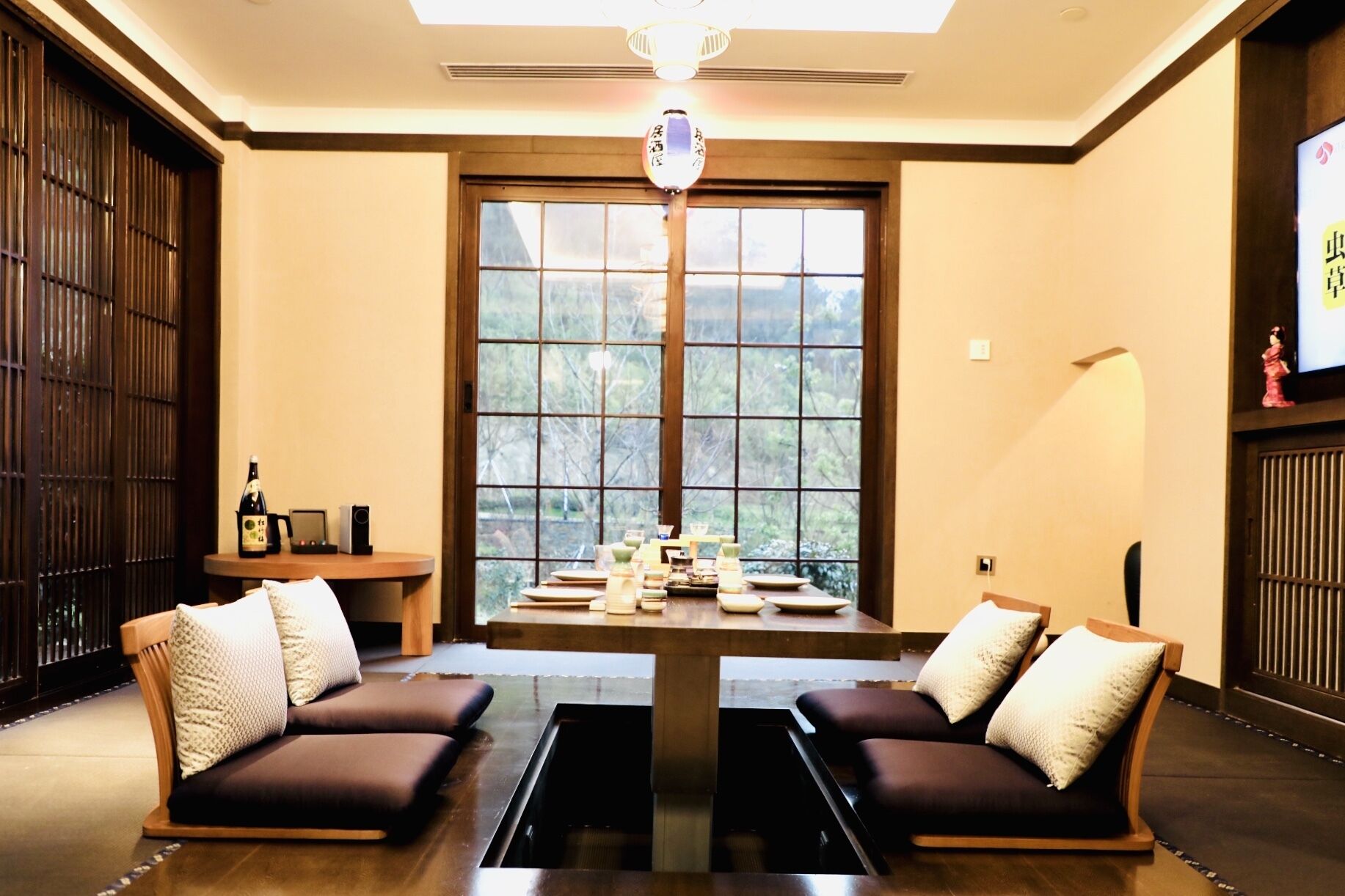 A room with two sofas facing a central table, a large window with a view of trees, and a traditional Japanese lantern.