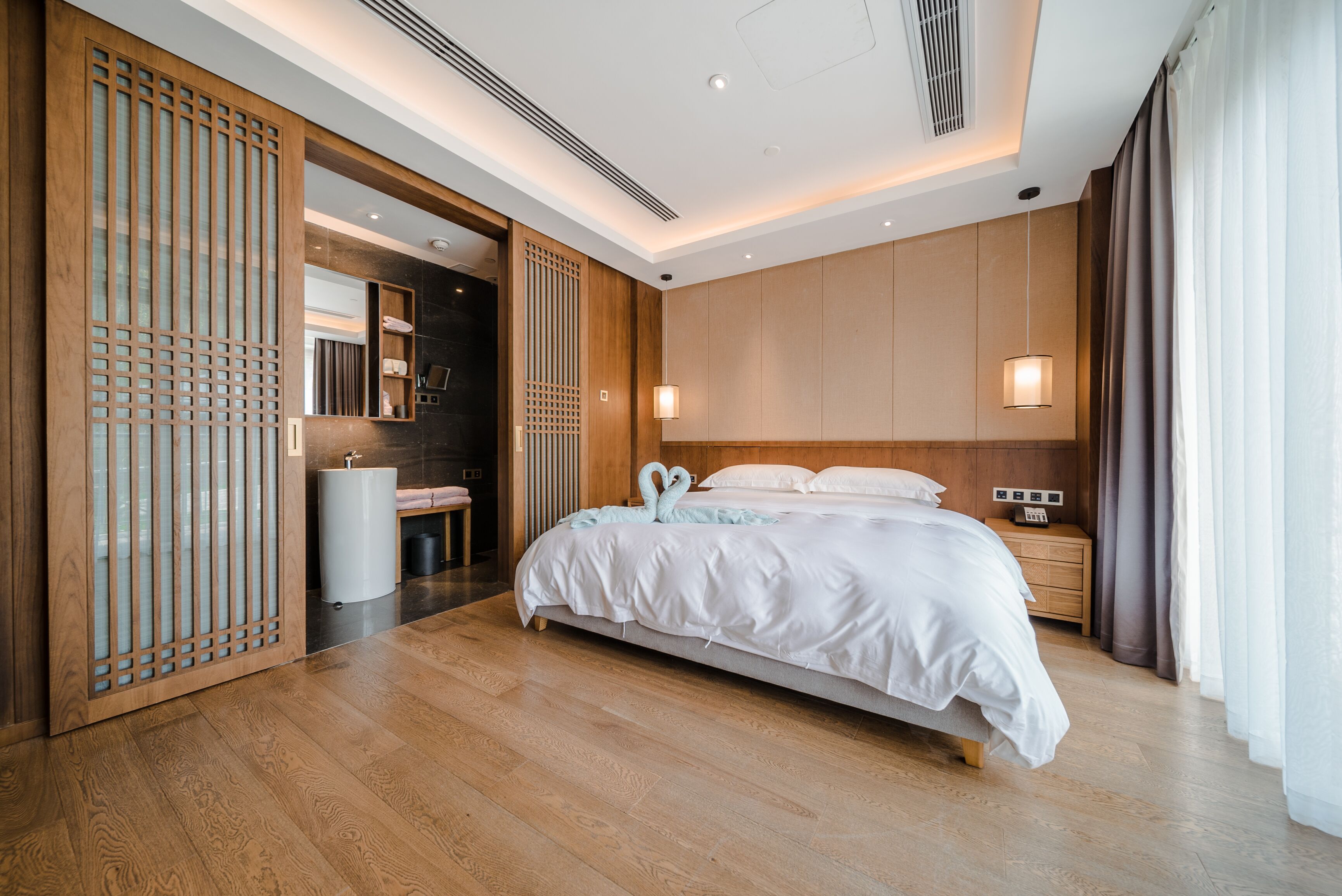 A modern hotel room with a wooden floor, a large bed with white bedding, a nightstand, a wall-mounted lamp, and a sliding door with a decorative screen.
