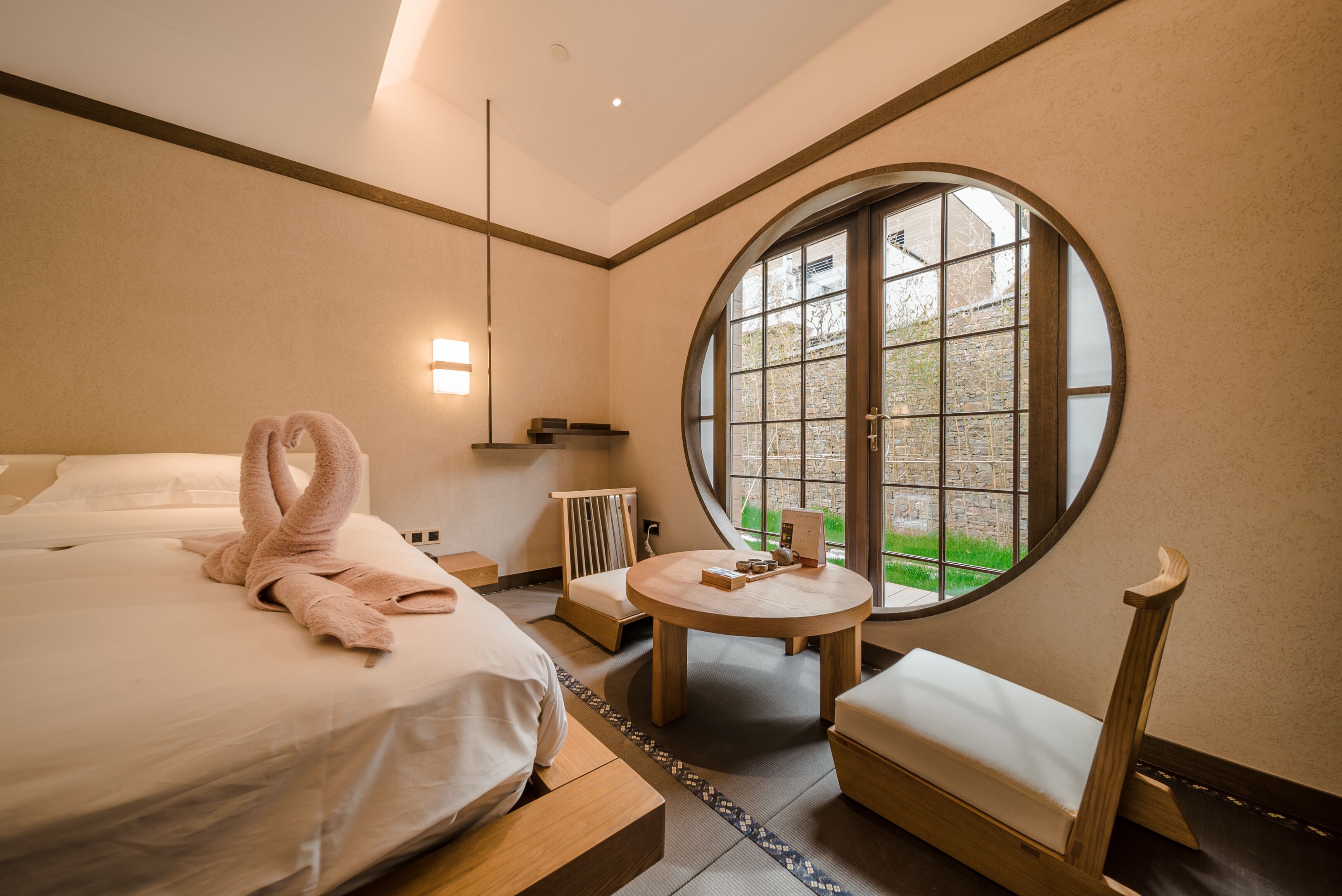 Habitación de hotel con cama, dos mesitas de noche, una mesa redonda, un banco y un ventanal con vistas a zonas verdes.