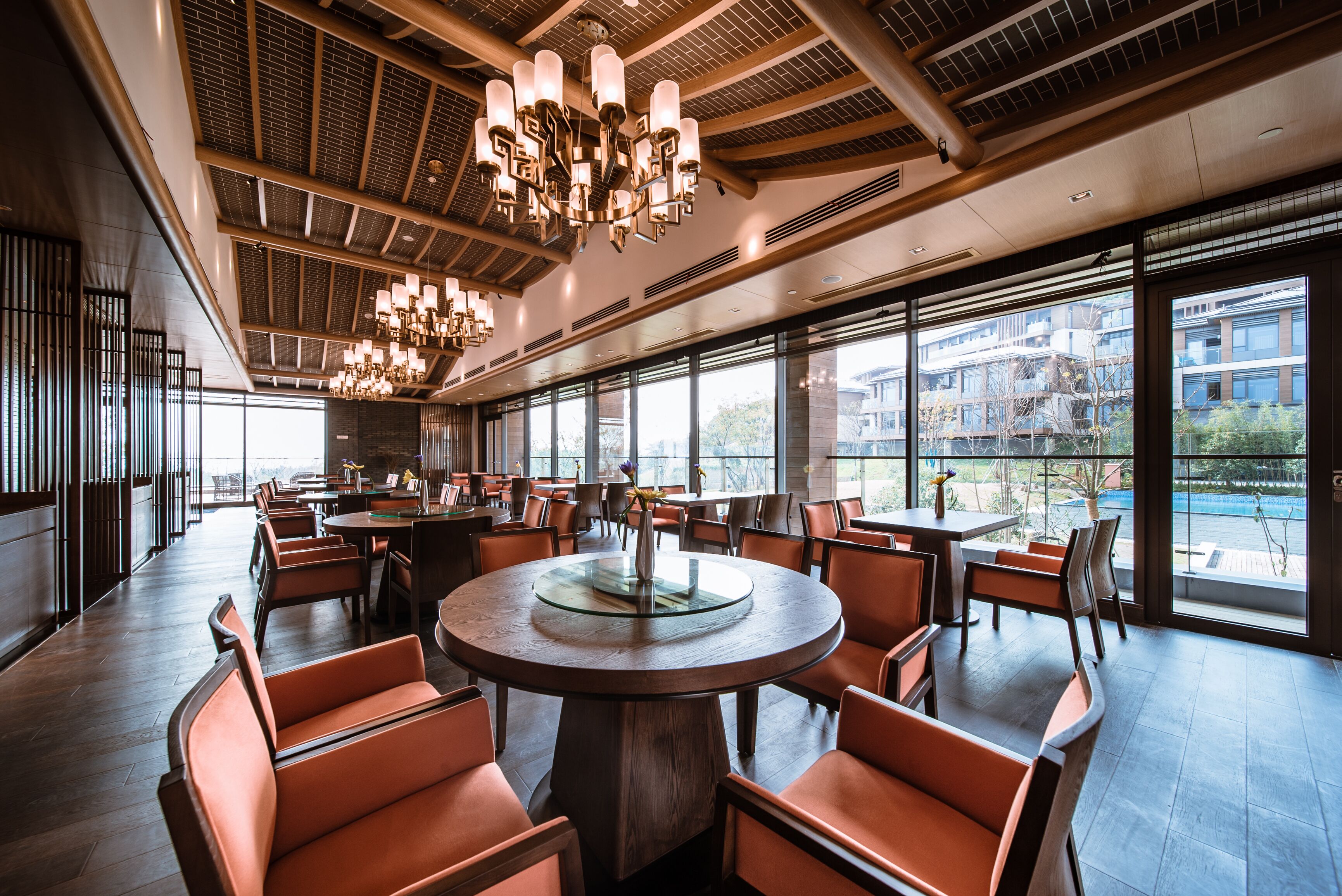 A spacious dining area with round tables and orange chairs, large windows offering views of buildings and a pool.