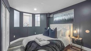 A bedroom with a large bed, two windows, a headboard with a light fixture, and a small bedside table with a lamp.