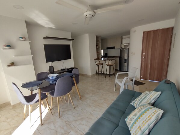 Ein Wohnzimmer mit einer Couch, einem Esstisch mit Stühlen, einem Fernseher und einer Küchenzeile mit Kühlschrank und Kaffeemaschine.