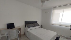 Un dormitorio con cama, escritorio con una computadora portátil, ventilador de techo y ventana.