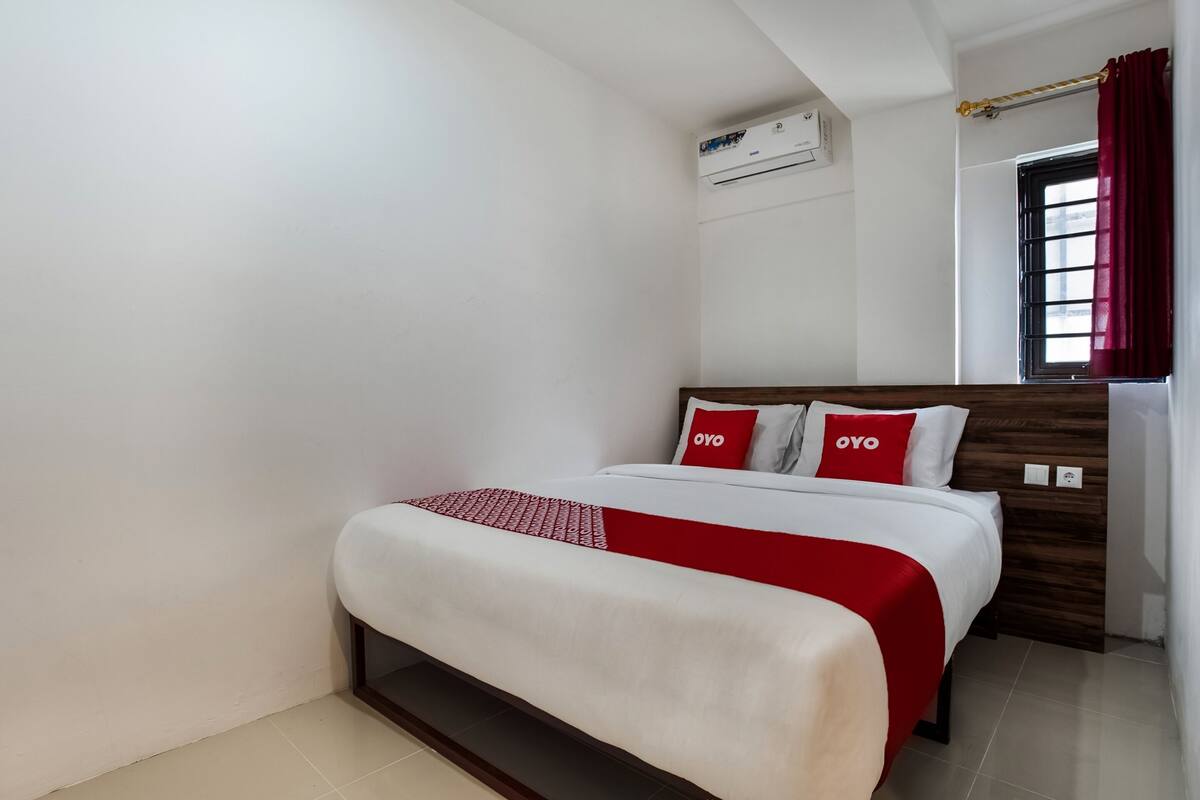 Una habitació d'hotel amb un llit, parets blanques, una finestra amb cortines vermelles i un aparell d'aire condicionat a la paret.