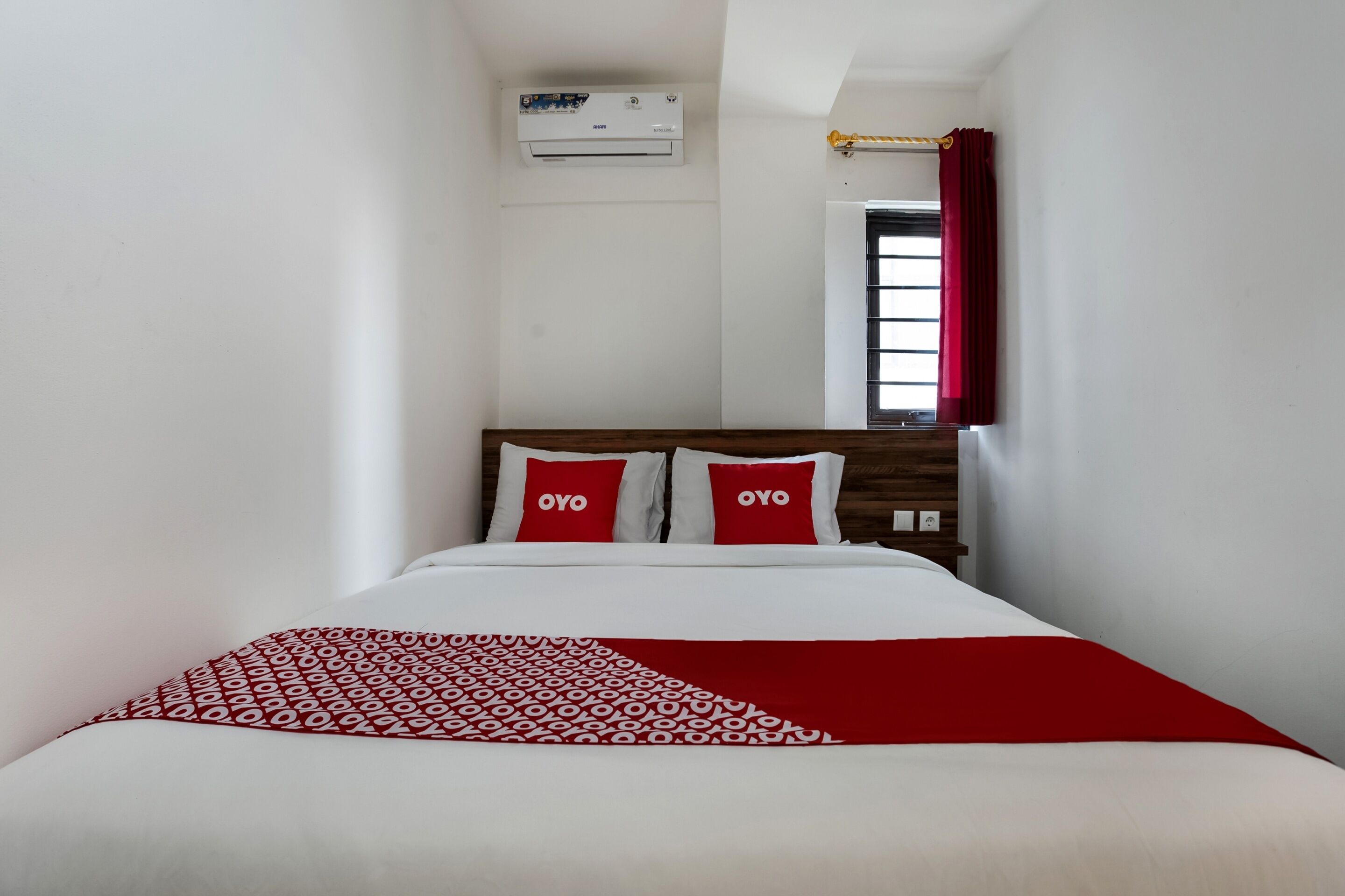 A hotel room with a bed, two pillows with 'OYO' branding, a red and white patterned throw, a window with red curtains, and an air conditioning unit on the wall.