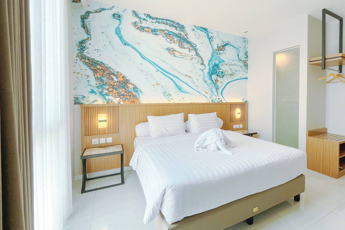 A hotel room with a large bed, a wooden headboard, a nightstand, a wall-mounted mirror, and a wooden wardrobe.