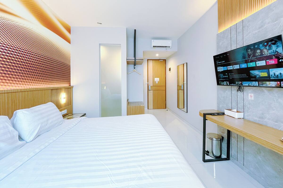 A modern hotel room with a large bed, a TV mounted on the wall displaying various channels, a wooden bedside table, and a small trash bin.