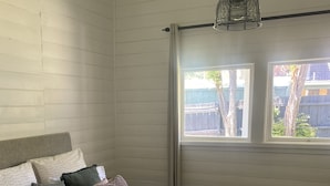 A bedroom with a bed, pillows, a blanket, a hanging light fixture, and a window with a view of trees and a fence. - Yarra Cottage (Yarra Junction)