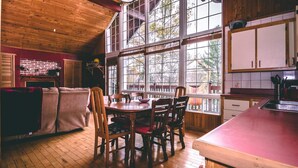 A cozy kitchen with wooden floors, a dining table, and large windows offering a view of trees. - Cozy cottage with hot tub! (Saint-Adolphe-d'Howard)