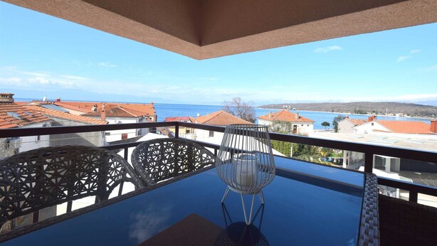 Uma varanda com mesa e cadeiras, dando para uma cidade costeira com edifícios de telhado vermelho e vista para o mar.