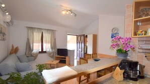 Uma sala de estar moderna com sofá, mesa de centro, TV e área de jantar com cadeiras. Há uma cozinha com cafeteira e pia.
