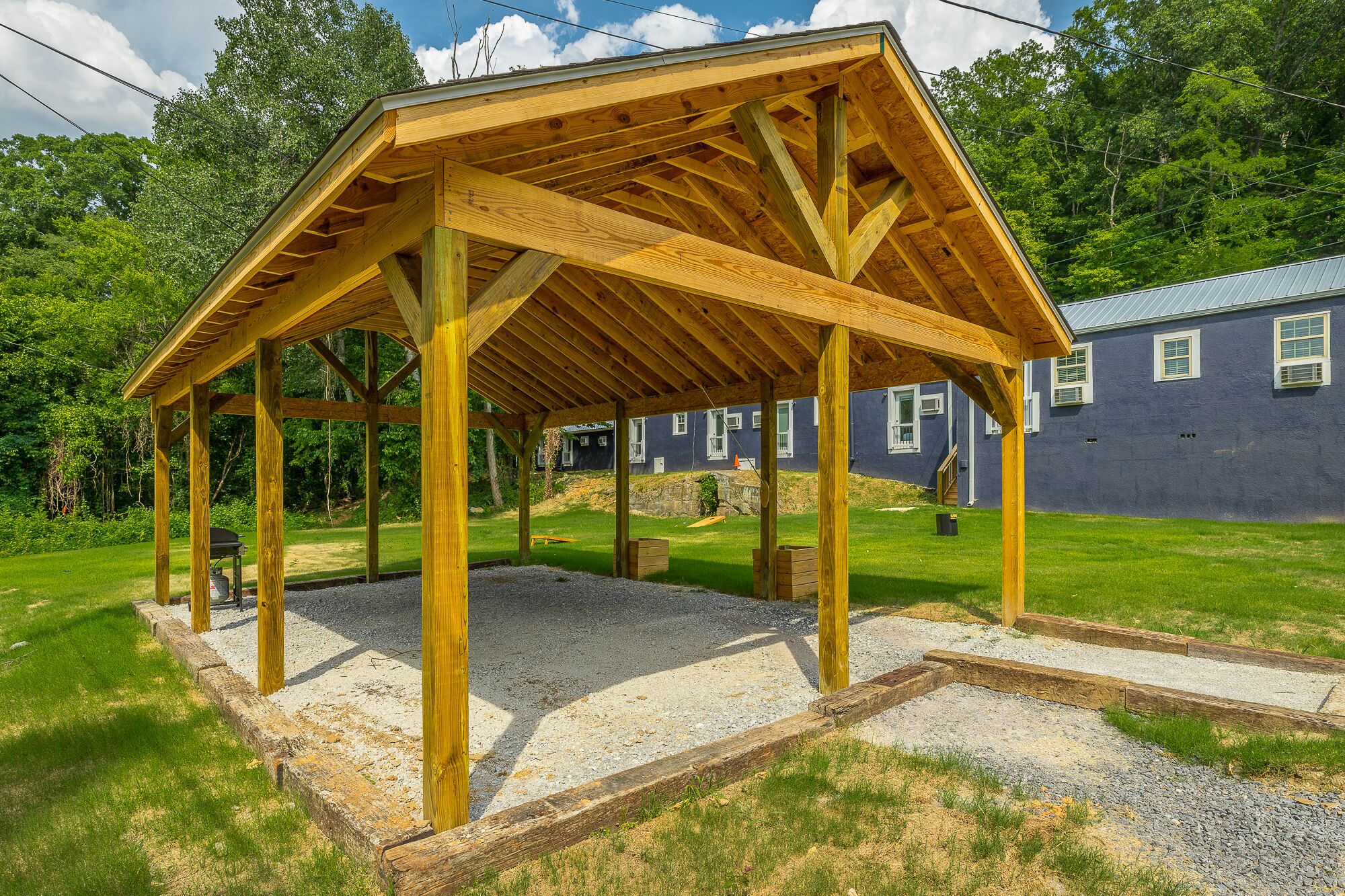 A wooden gazebo structure with a gravel base, surrounded by a grassy area and a house in the background.
