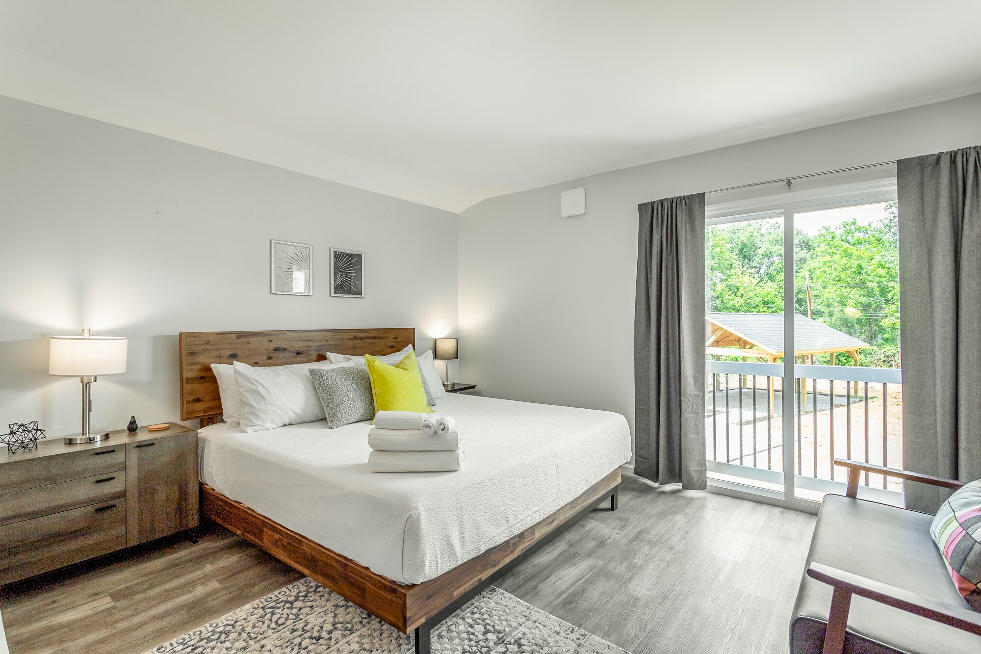 A hotel room with a wooden bed, a nightstand, a desk, and a view of a balcony with a wooden structure.
