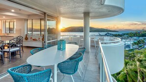 Un balcon avec une table blanche et des chaises bleues, offrant une vue panoramique sur un plan d’eau et des collines au coucher du soleil.