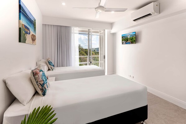 A modern hotel room with two beds, a ceiling fan, an air conditioning unit, and a large window offering a view of the outdoors.