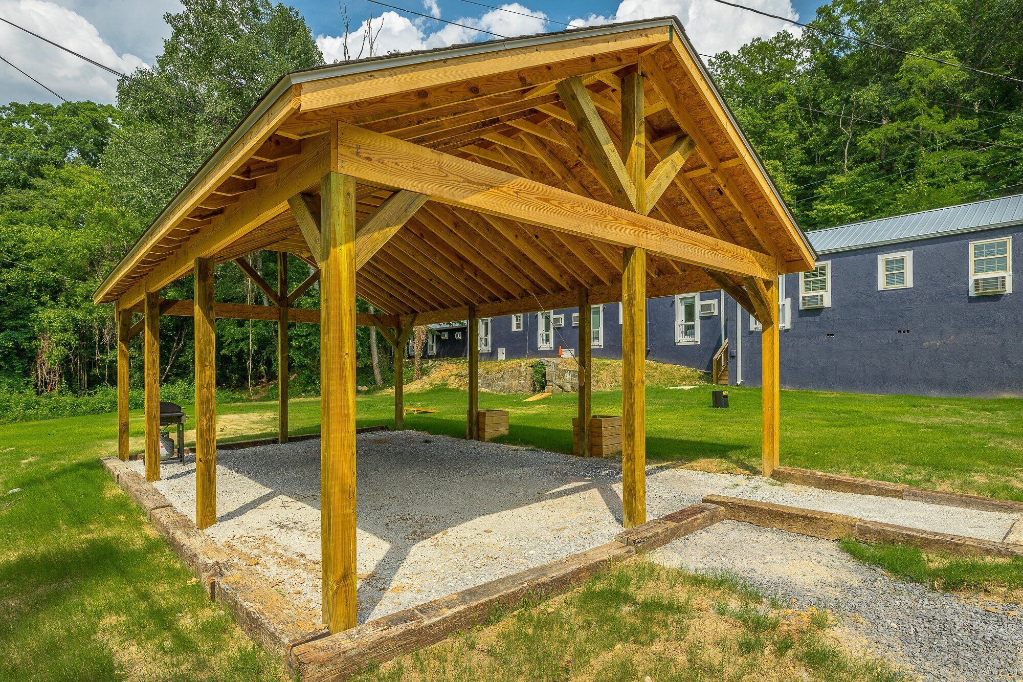 A wooden gazebo structure with a gravel base, surrounded by a grassy area and a house in the background.