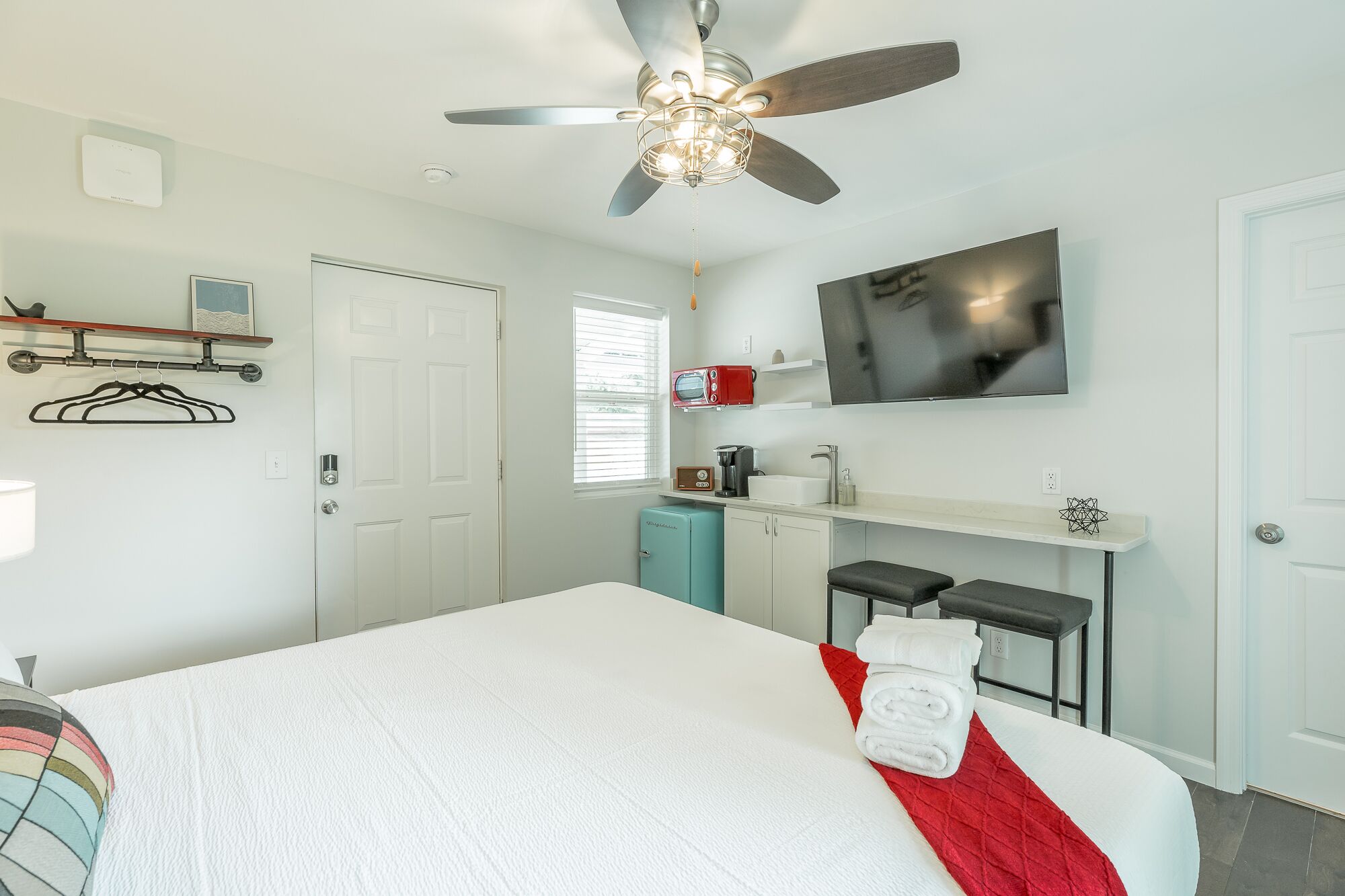 A hotel room with a bed, a ceiling fan, a TV, a small kitchenette, and a window.