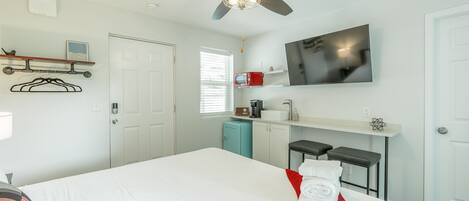 A hotel room with a bed, a ceiling fan, a TV, a small kitchenette, and a window.