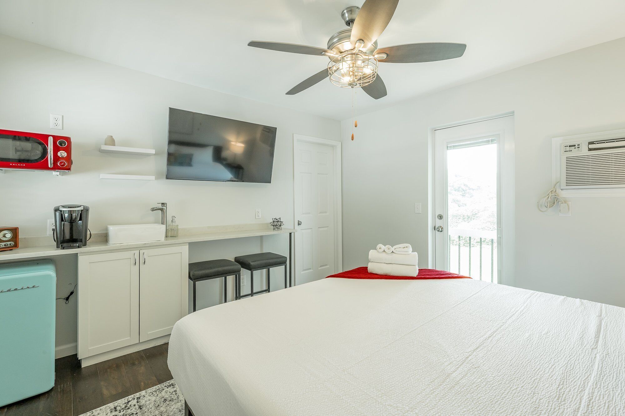Une chambre comprenant un lit, un ventilateur de plafond, une télévision, un coin cuisine et une porte donnant sur un balcon.