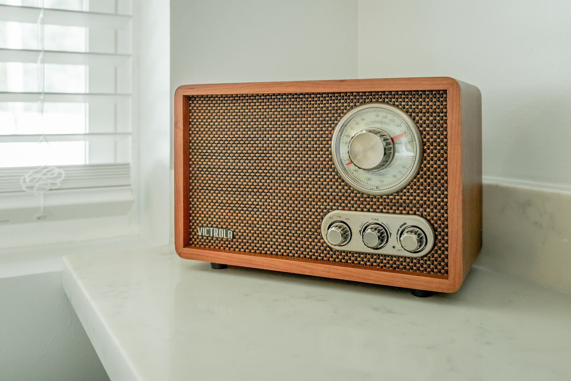 Una radio d'epoca con cornice in legno e griglia frontale, posata su una superficie bianca vicino a una finestra con persiane.