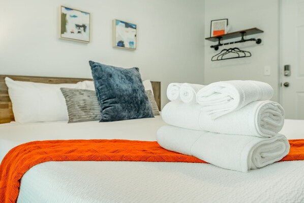 A neatly made bed with white linens and rolled towels, a blue pillow, and an orange throw blanket.