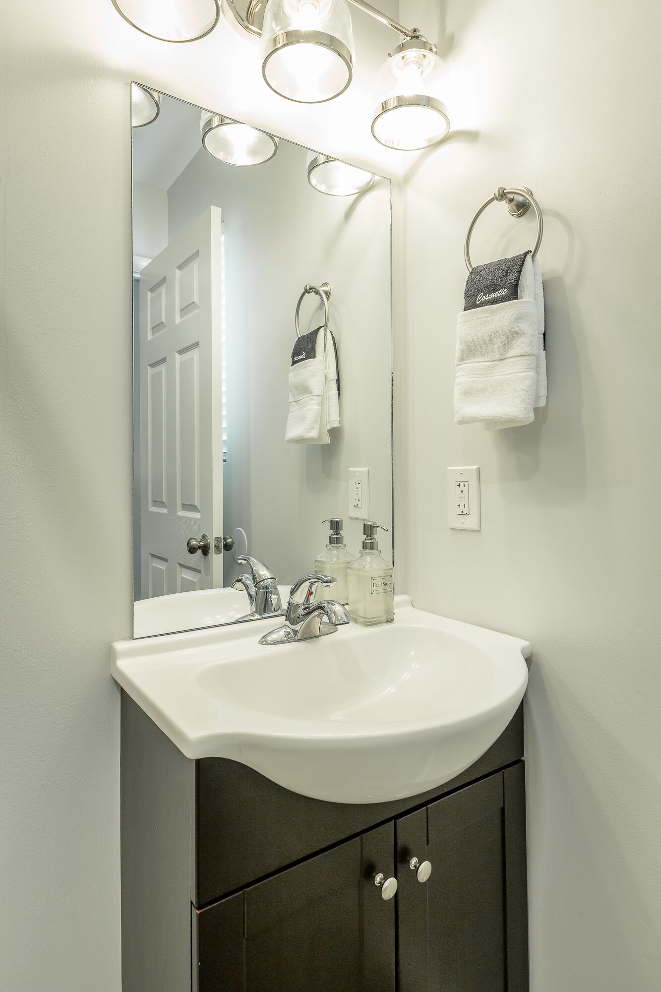 A bathroom with a white sink, two faucets, a mirror with lights, and a towel rack with a white towel and a gray towel.