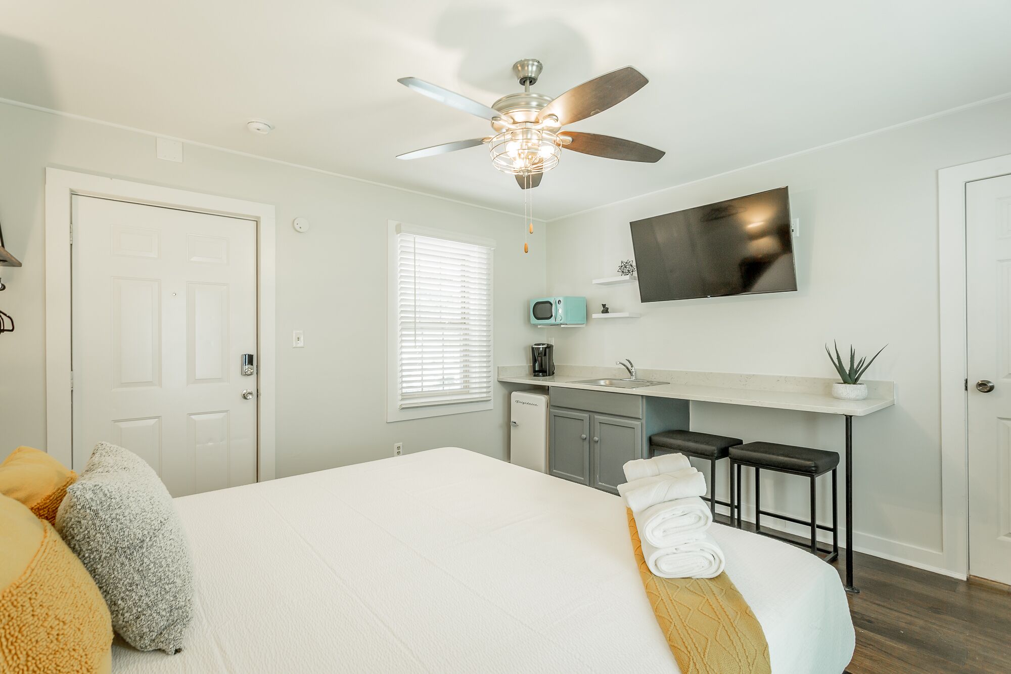 A hotel room with a bed, a ceiling fan, a TV, a kitchenette, and a bathroom.