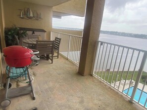 A balcony with a view of a body of water, a grill, a table, and chairs.