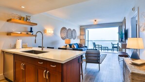 A modern kitchen with a large island, wooden cabinets, and built-in shelves. There is a dining area with a sofa and chairs, and a large window offering a view.