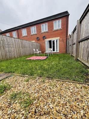 A brick house with a wooden fence, a grassy yard, and a patio with chairs.