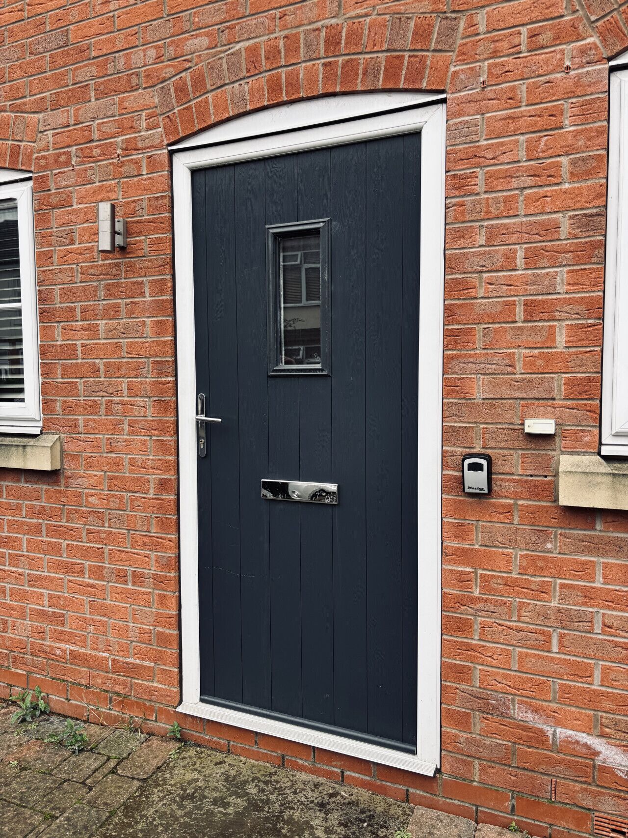 A dark-colored door with a small window and a handle, set in a brick wall with a white trim.