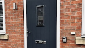 A dark-colored door with a small window and a handle, set in a brick wall with a white trim.