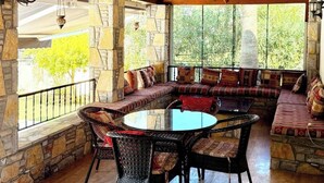 Une terrasse couverte avec des murs en pierre, une table ronde en verre, des chaises en osier et un banc intégré avec des coussins.