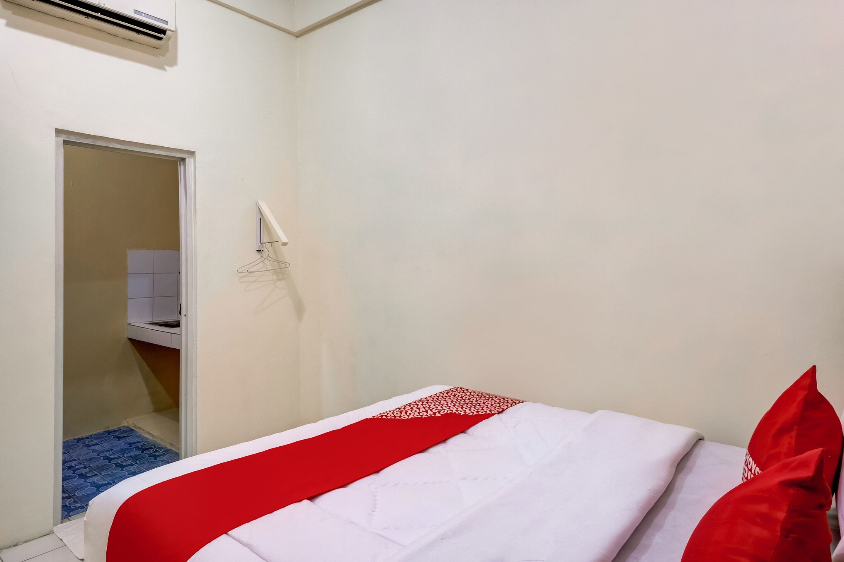 Ein Hotelzimmer mit einem Bett, einer roten und weißen Bettdecke, einem roten Kopfkissen, einer weißen Wand, einer Tür zu einem Badezimmer und einer Klimaanlage.