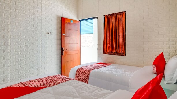 Een hotelkamer met twee bedden, wit beddengoed, rode accenten, een muur met een baksteenmotief, een raam met gordijnen en een houten deur.
