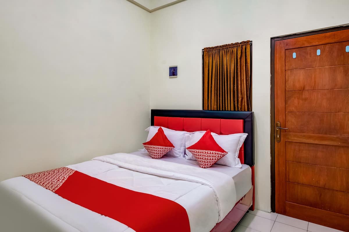 Een net opgemaakt bed met rood-witte beddengoed, een houten deur en een klein, aan de muur bevestigd lampje.