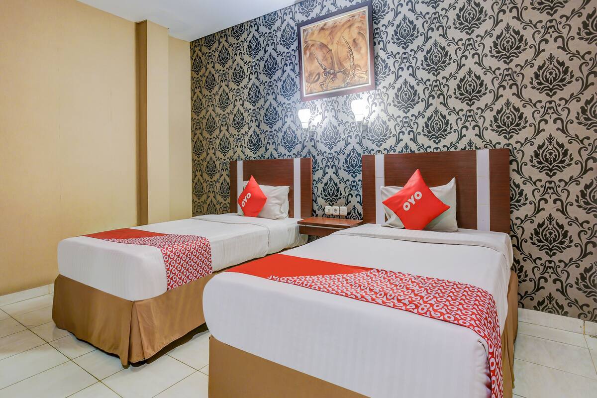 Een hotelkamer met twee bedden, elk voorzien van een rood-wit gebeddeken, een houten hoofdbord en een rode kussen met het logo "oyo". De muren zijn bekleed met blauw-zwart behang en erboven hangt een kunstwerk in een lijst.