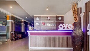 A reception desk with a modern design, featuring the name 'TRISULA' prominently displayed. There are clocks above the desk, and a large vase with decorative sticks to the right. - OYO 3936 Hotel Trisula Makassar (Makassar)