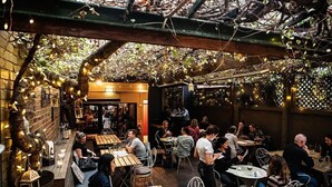 An outdoor dining area with wooden tables and chairs, surrounded by a wooden pergola and string lights.