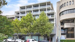 A multi-story building with balconies, parked cars, and trees in front.