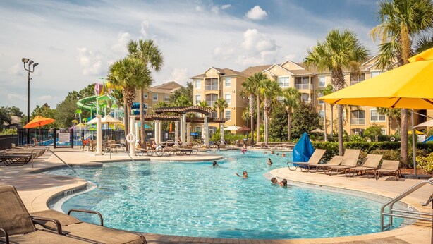 A swimming pool area with lounge chairs and umbrellas, surrounded by palm trees and a building with balconies. - The Wish House, just miles from Disney World (Four Corners)