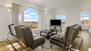 A modern living room with a large TV, leather sofas, and a view of a cityscape through large windows. - Breathtaking View 2BD/1BTH Unit in Central SD (San Diego)