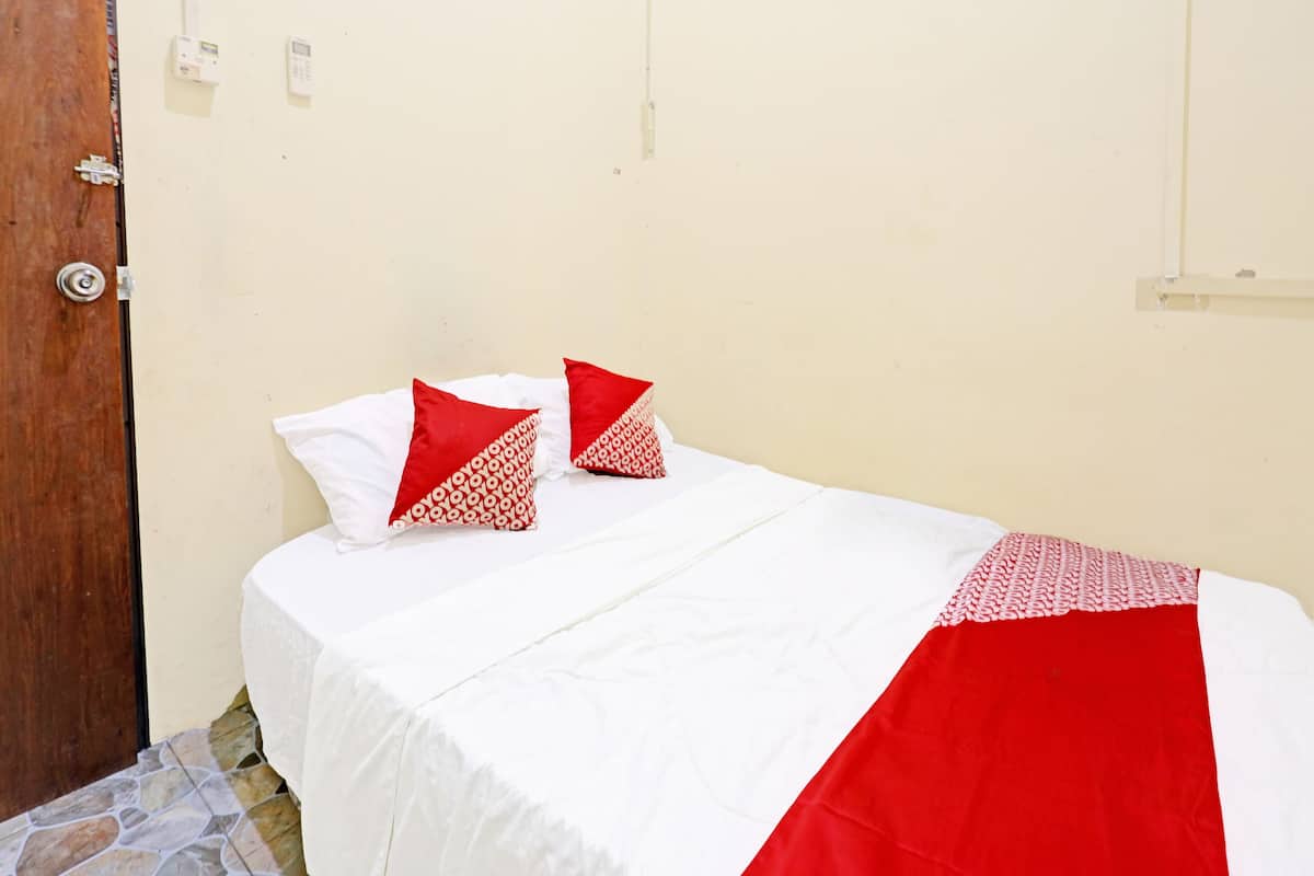 A single bed with white bedding and red accents, a wooden door, and a plain wall with a light switch.
