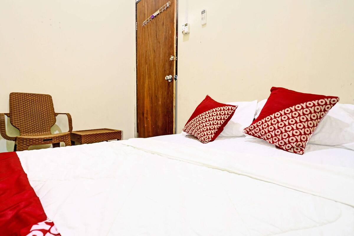 A bed with white bedding and red pillows, a wicker chair, and a wooden door.
