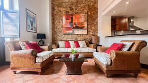A living room with wicker furniture, a stone wall, and a kitchen in the background.